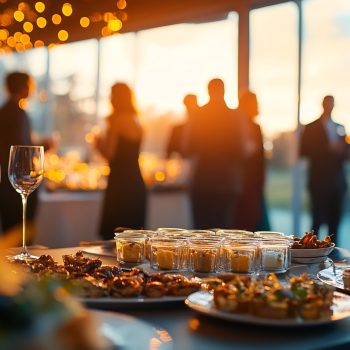 Elegant buffet table with gourmet appetizers and wine glasses at a sunset outdoor event. People socializing at a formal gathering